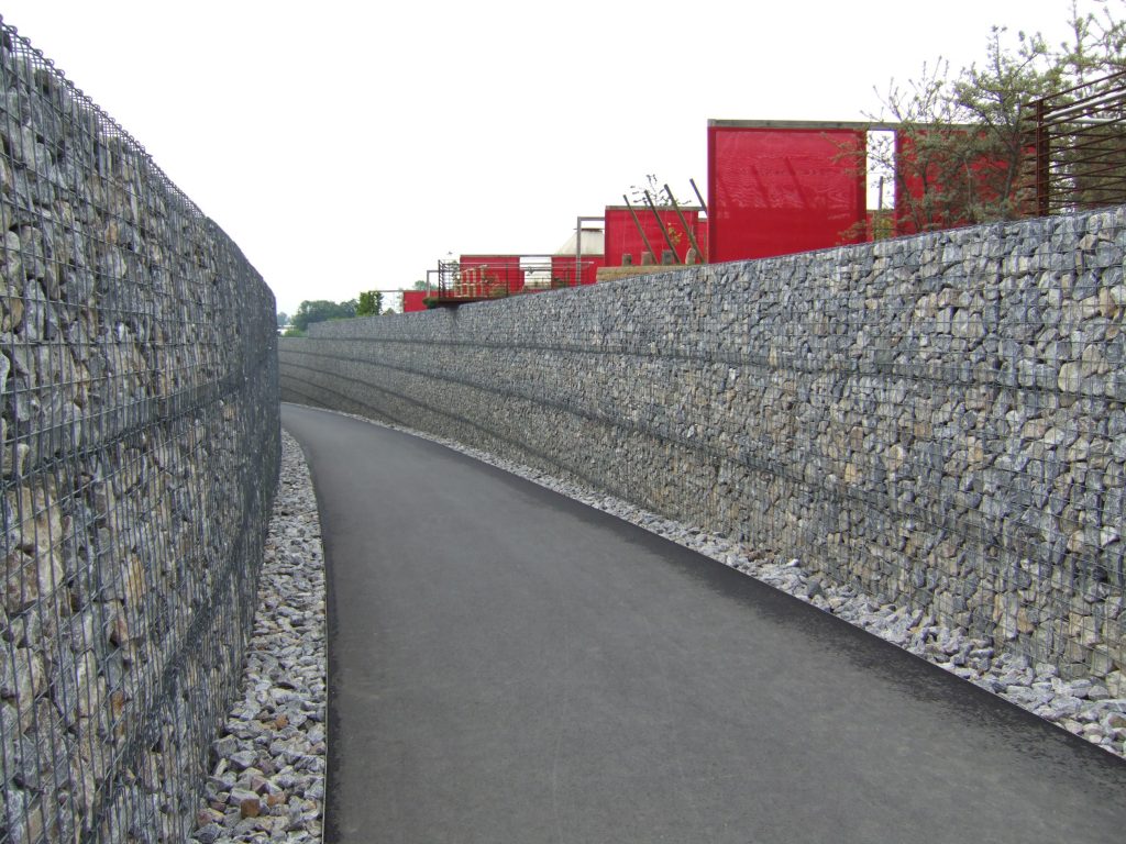 Asphaltierter Weg zwischen hohen Stützmauern aus Steingabionen mit moderner Architektur im Hintergrund.