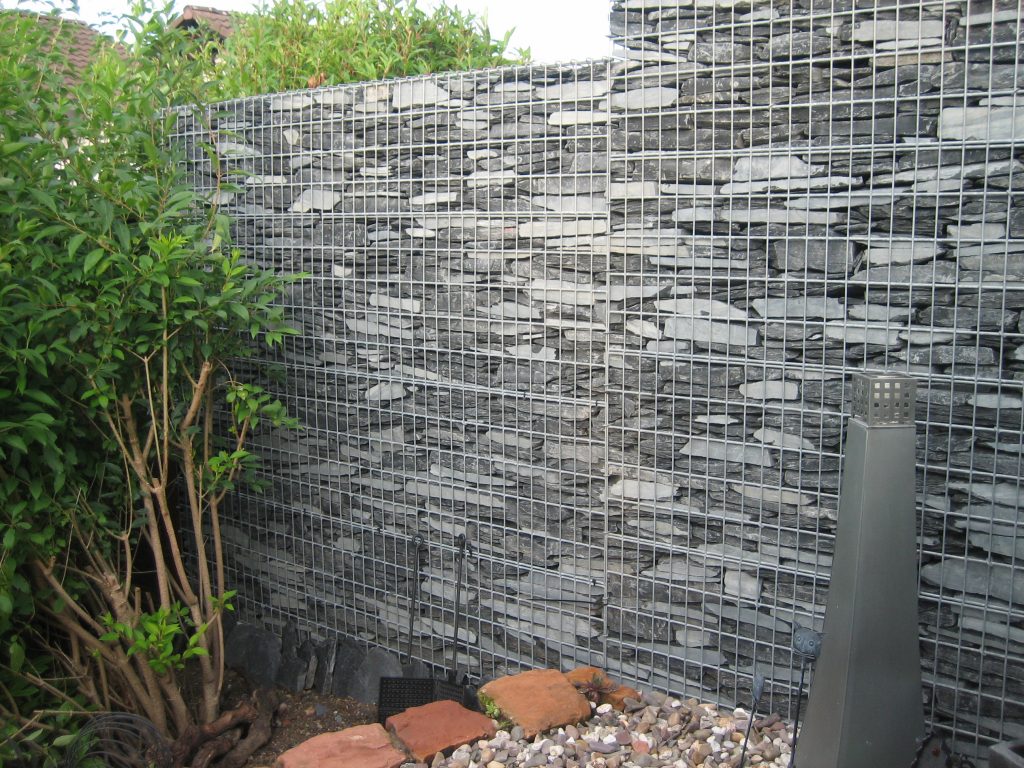 Moderne Sichtschutzwand aus dunklen Schiefergabionen im Garten, umgeben von Pflanzen und Kiesdeko.