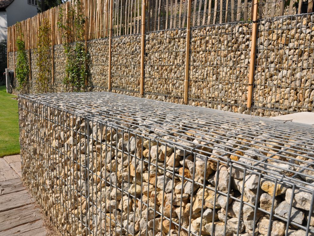 Moderne Gartenmauer aus hellen Steingabionen mit Holzelementen und Rankpflanzen als Sichtschutz.