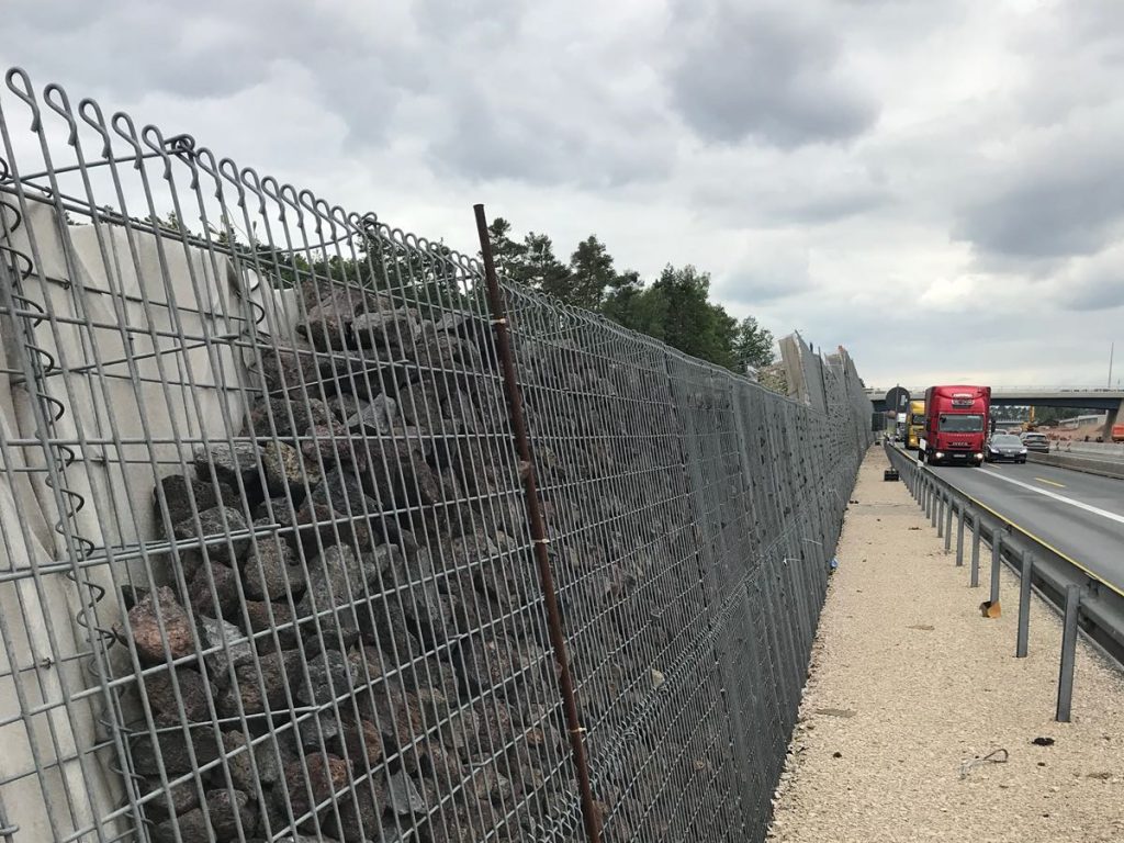 Lärmschutzwand aus Steingabionen entlang einer Autobahn mit vorbeifahrenden Lastwagen.