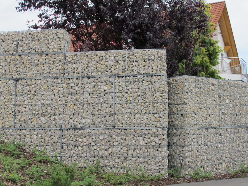 Mehrstufige Stützmauer aus Steingabionen als Hangbefestigung vor einem Wohnhaus mit Garten.