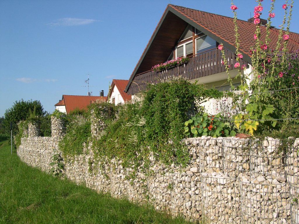 Steingabionen mit üppiger Bepflanzung als Gartenmauer vor einem Einfamilienhaus mit rotem Dach.